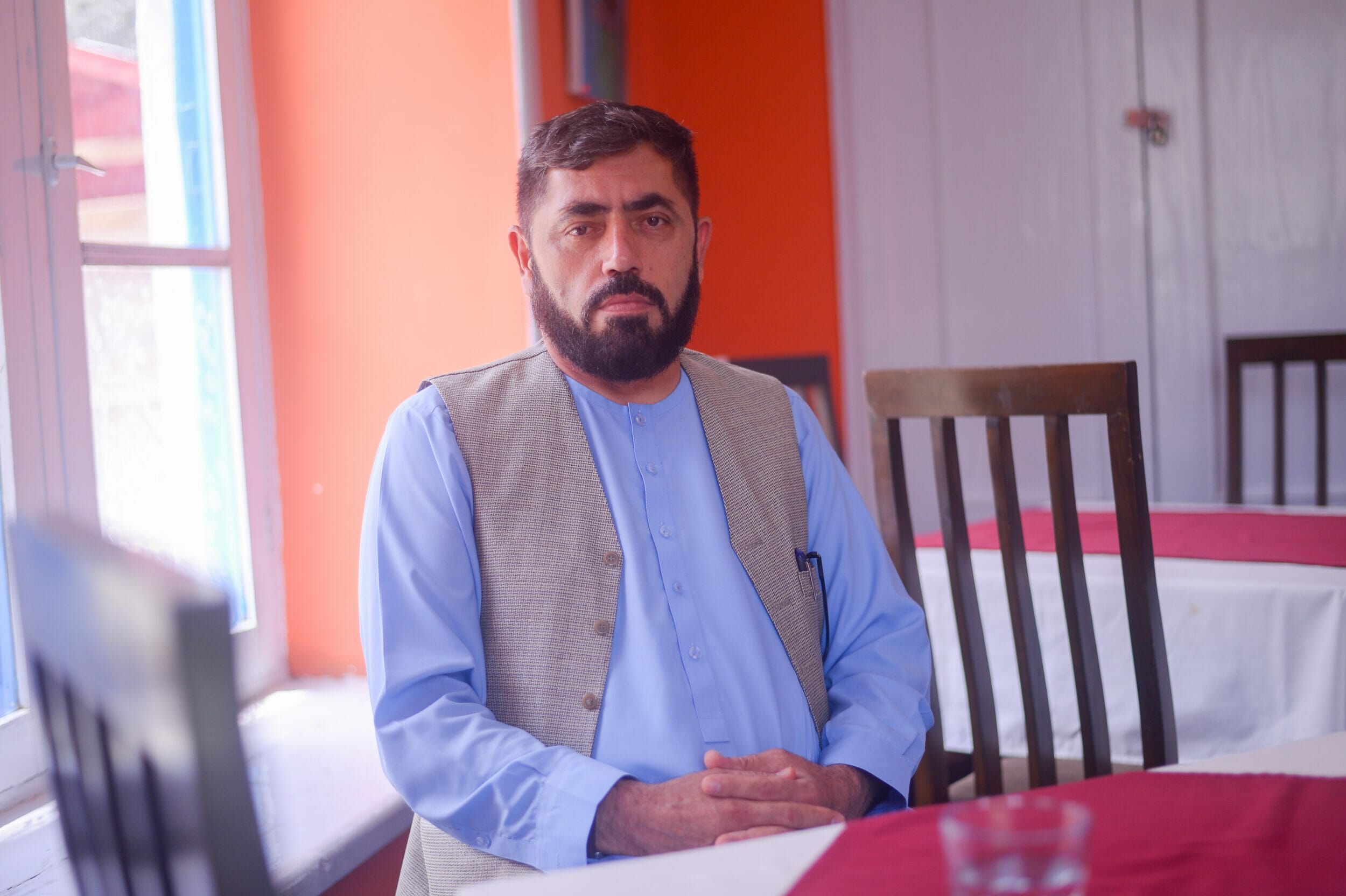 Yasin Adel sitzt mit tradioneller, blauer afghanischer Kleidung und grauer Weste vor einem roten Tisch. Er trägt kurze Haare und einen leichten Vollbart.