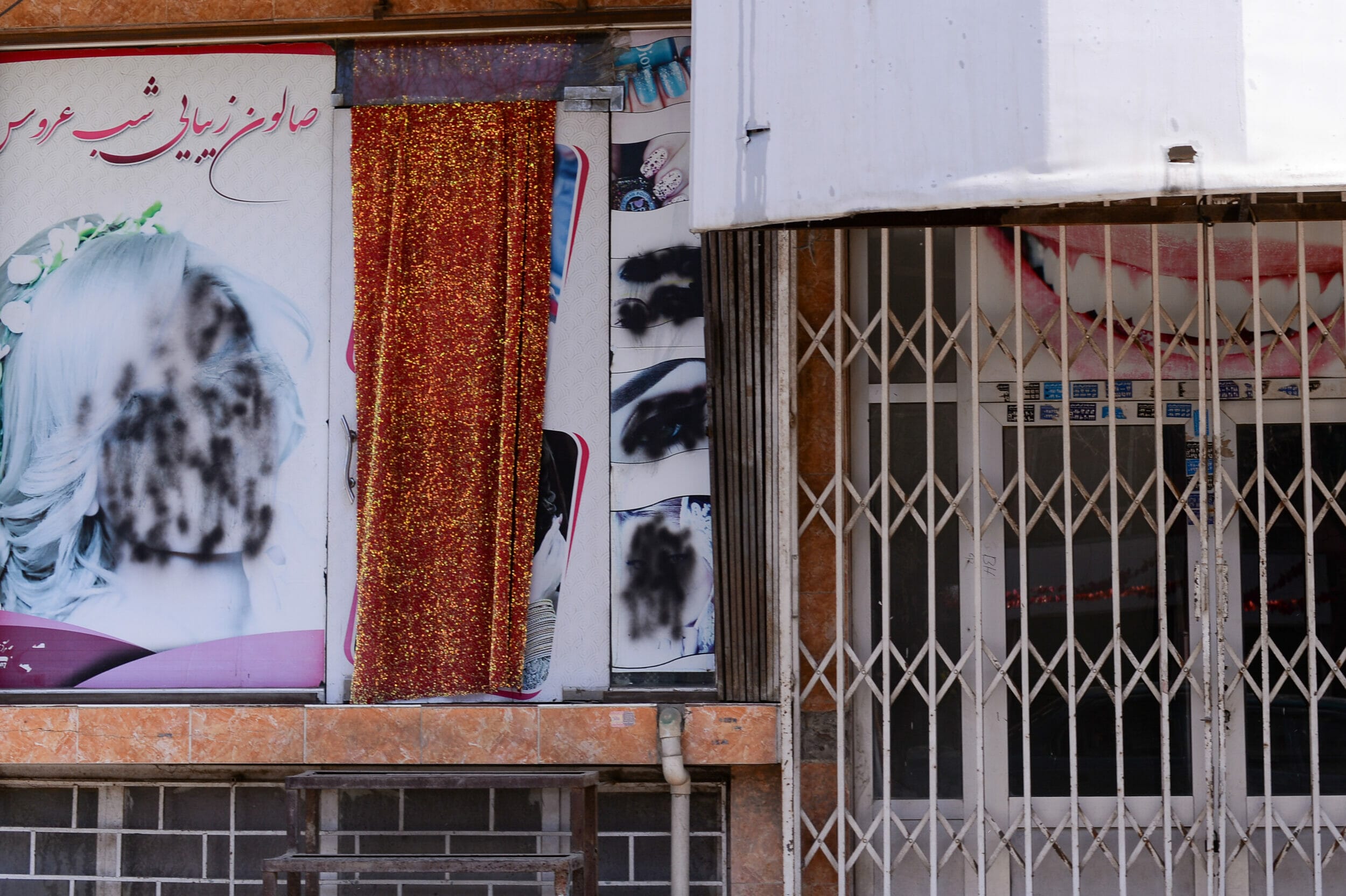 Foto von Werbung für einen Friseursalon, auf dem Frauengesichter mit schwarzer Farbe übermalt sind. Daneben ein geschlossenes Gittertor.