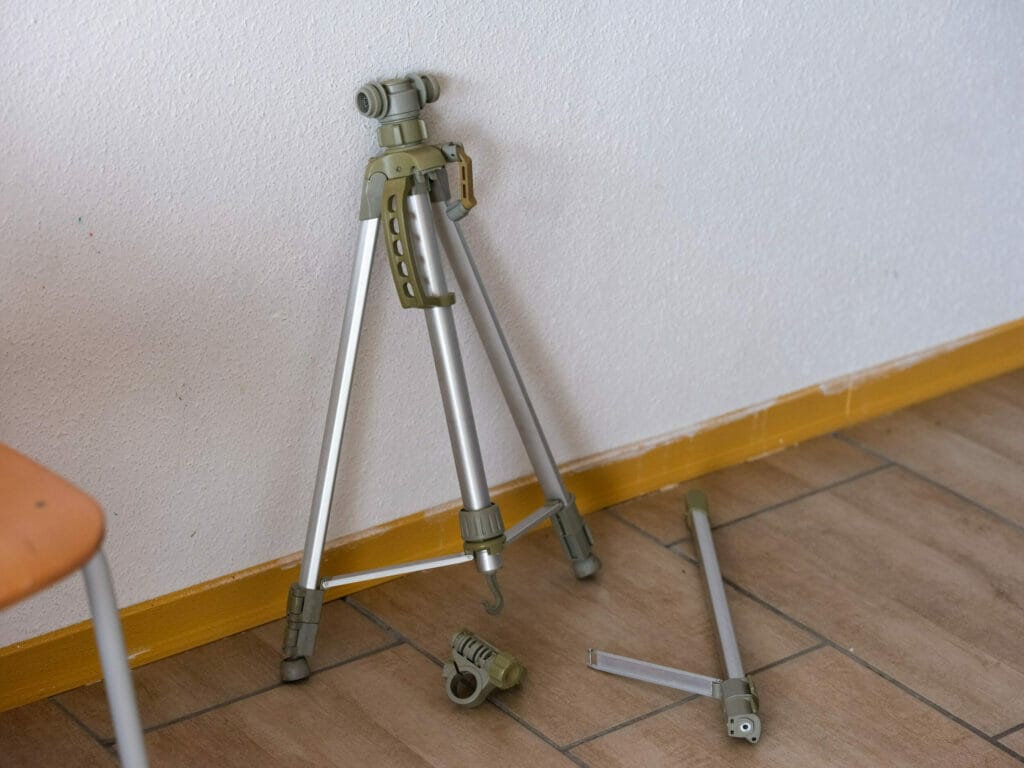 A camera tripod on the floor next to a chair has obviously suffered damage: one of its struts has broken off.