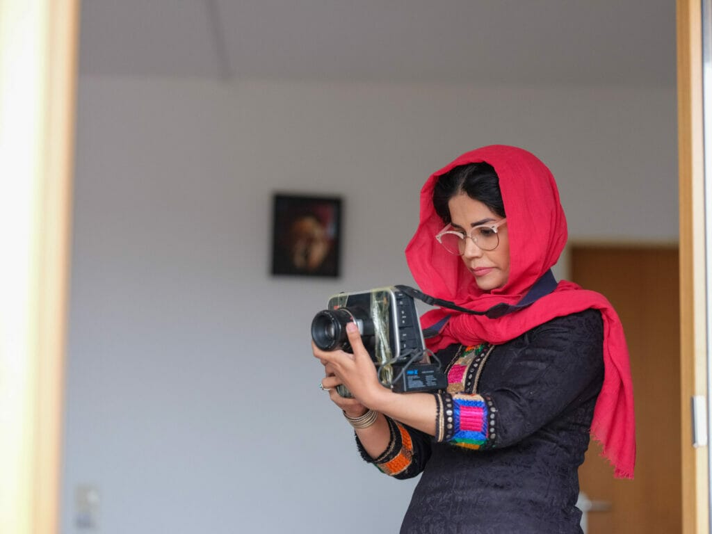 A young woman wears a red headscarf with dark curly hair peeking out from under it, along with a traditional Afghan robe with a black base color and red-pink-purple decorations on the sleeves. She is seen in profile and looks intently at the camera she is holding in front of her.  In the background, a small picture hangs on the wall, which can only be seen out of focus. Zainab Entezar is one of the first Afghans to be evacuated to Germany through the Federal Reception Program.