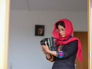 Eine junge Frau trägt ein rotes Kopftuch, unter dem dunkle lockige Haare hervorlugen, dazu ein traditionell afghanisches Gewand mit schwarzer Grundfarbe und rot-pink-violetten Verzierungen an den Ärmeln. Sie ist im Profil zu sehen und schaut konzentriert auf die Kamera, die sie vor sich hält. Im Hintergrund hängt ein kleines Bild an der Wand, das nur unscharf zu sehen ist.