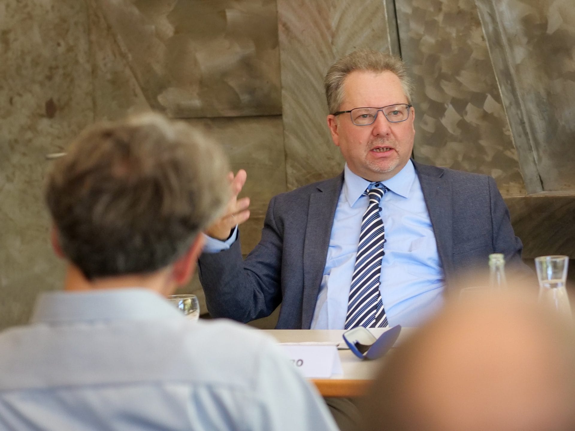 Shlomo Shpiro sitzt am Tisch und hebt die rechte Hand.