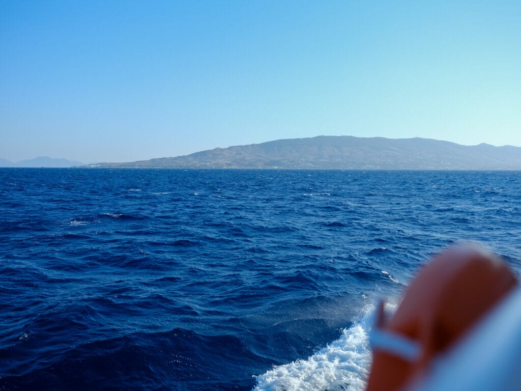 Ein Blick auf das Meer von einem Boot aus, eine Insel türmt sich im Hintergrund auf. Das Meerwasser ist tiefblau und wellig.