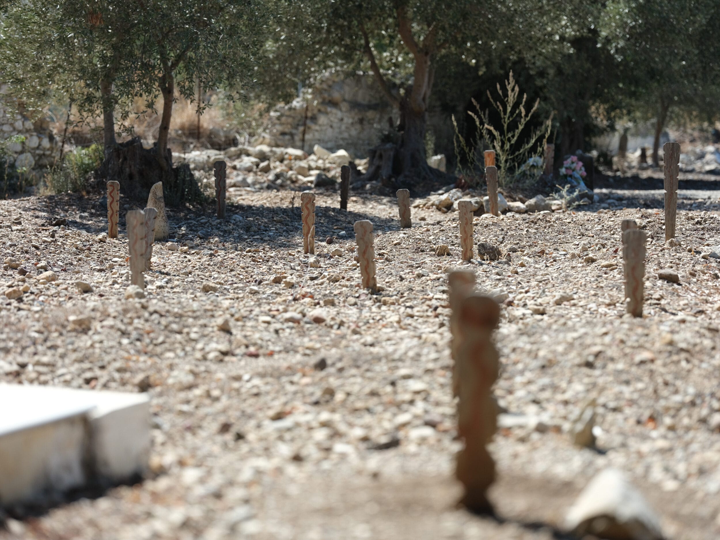 Bodensperspektive anonymer islamischer Gräber auf Kos: geschwungene Hölzer mit einer rötlichen Wellenlinie stecken in einem kieslastigen staubigen Boden, es gibt keine sichtbaren Grabhügel oder befestigte Gräber.