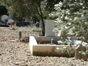 Ein Friedhof mit einem Grabstein inmitten eines Erdfeldes, im Hintergrund anonyme Holzmarkierungen muslimischer Gräber.