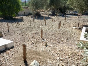 Ein mit Steinen bedeckter staubiger heller Boden, aus dem einzelne geschwungene Holzpfeile ragen, die muslimische anonyme Gräber kennzeichnen. Im Hintergrund ein paar blassgrüne Bäume, links und rechts angeschnitten weiße Grabumrandungen.