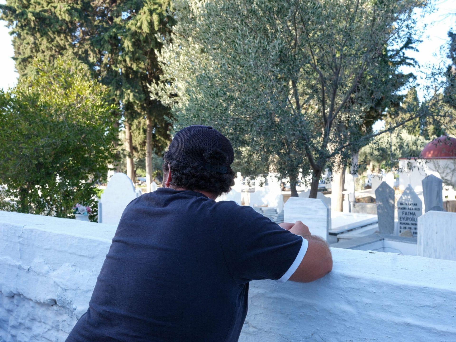 Ein Mann mit lockigen schwarzen Haaren, die unter einer Cappy hervorquellen von hinten mit abgewandtem Gesicht fotografiert, an einer weißen Mauer lehnend, hinter der Grabsteine sichtbar sind.