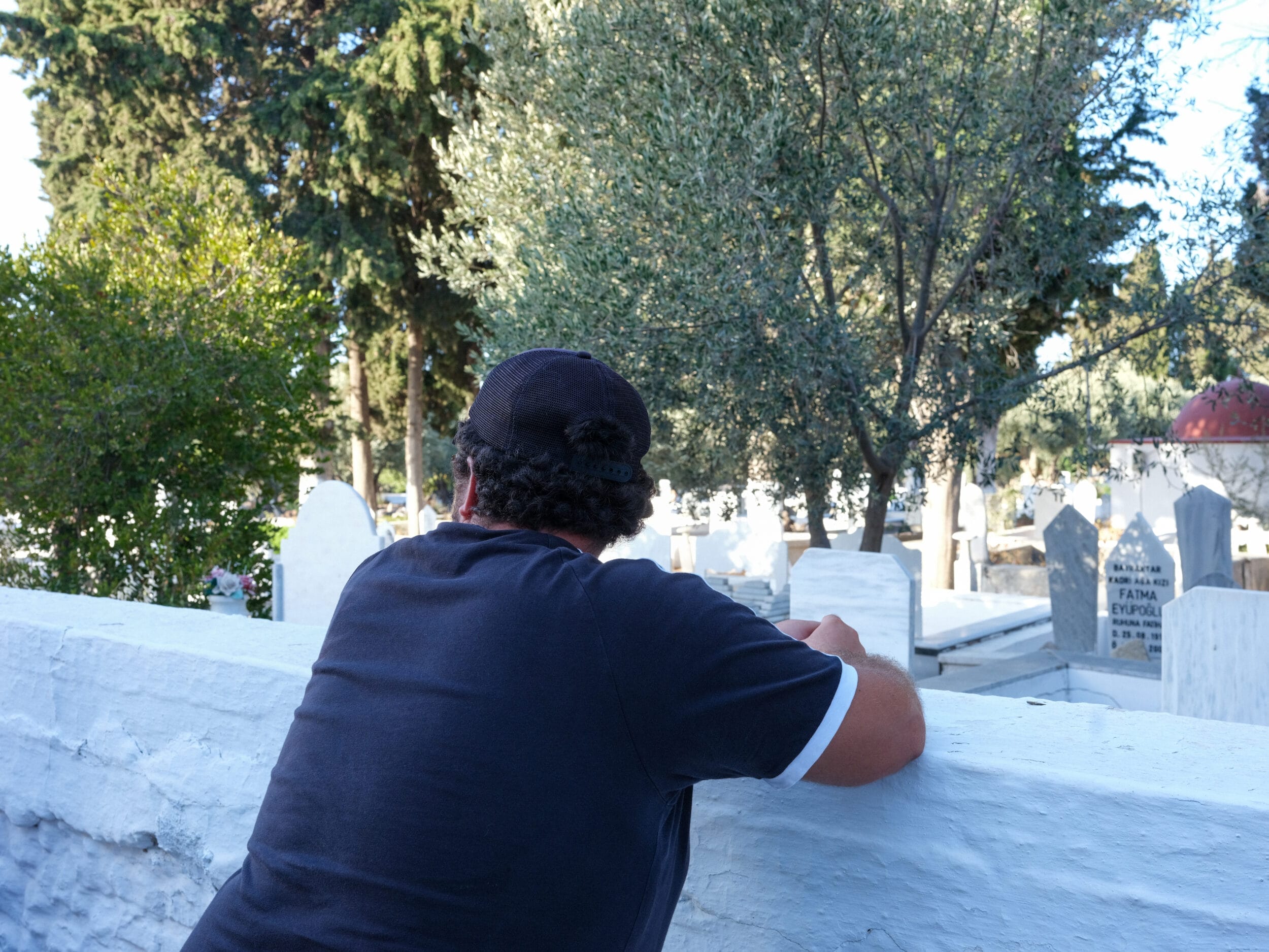 رجل ذو شعر أسود مجعد تم تصويره من الخلف ووجهه متجنب، متكئا على جدار أبيض تظهر خلفه شواهد القبور.