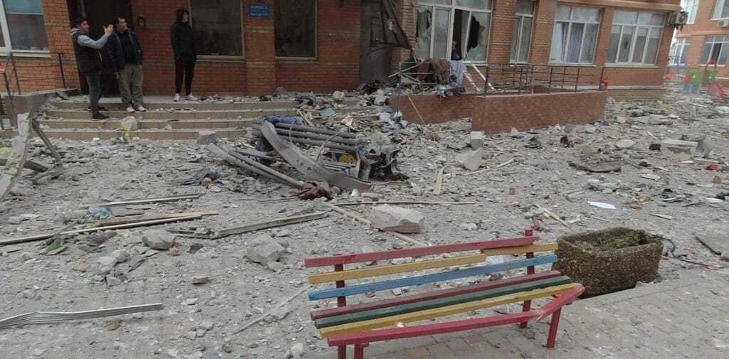 Debris and damaged infrastructure in front of a building, with onlookers present.