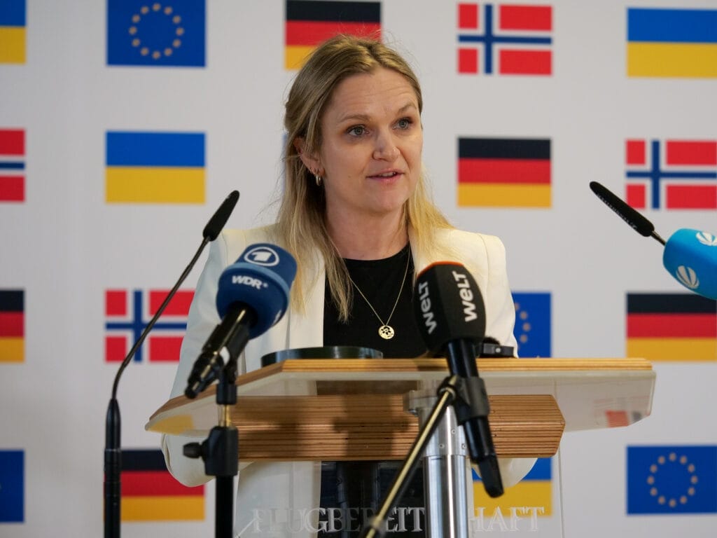 Eine Frau spricht an einem Podium mit Mikrofonen und einem Hintergrund aus EU-Flaggen.