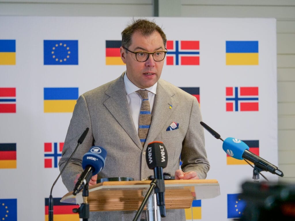 Ein Mann im Anzug spricht auf einem Podium mit Mikrofonen und Flaggen europäischer Länder im Hintergrund.