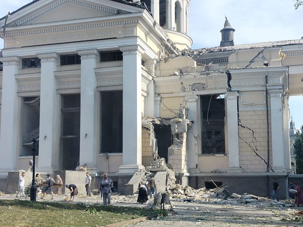 People gather around a heavily damaged classical-style building with debris strewn around.