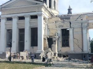 People gather around a heavily damaged classical-style building with debris strewn around.