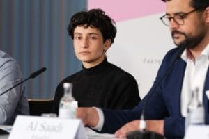 Podiumsdiskussion auf einer Konferenz mit einer konzentrierten Frau in einem schwarzen Rollkragenpullover und kurzen lockigen Haaren, die zwischen zwei männlichen Teilnehmern sitzt.
