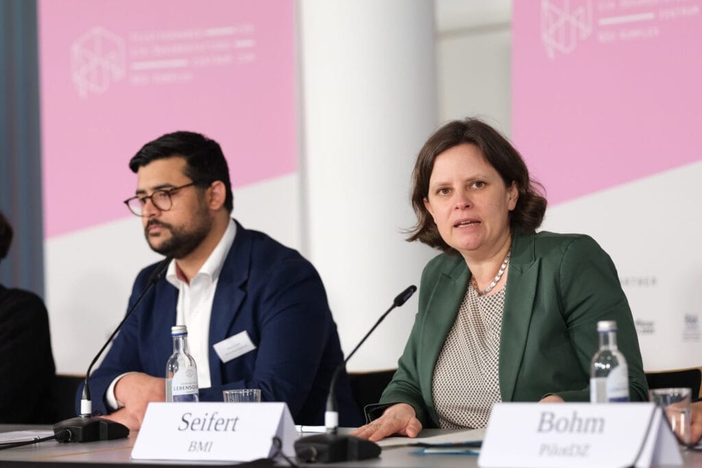 Zwei Diskussionsteilnehmer, eine Frau und ein Mann, diskutieren während einer Konferenz, mit Namensschildern und Mikrofonen vor sich.