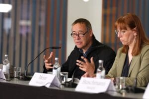 Two panelists, a man and a woman, were discussing at a conference table with name tags and bottles of water in front of them.