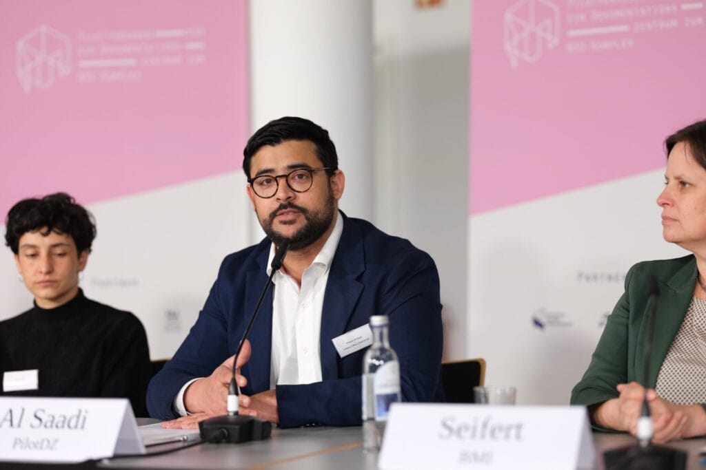 Ein Mann im blauen Anzug spricht bei einer Podiumsdiskussion, flankiert von zwei weiteren Teilnehmern, in einem Raum mit Plakaten mit Logos und Namen.