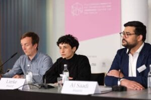 Drei Diskussionsteilnehmer einer Konferenz sitzen mit Namensschildern hinter Mikrofonen und diskutieren in einem Raum mit einem Banner im Hintergrund.