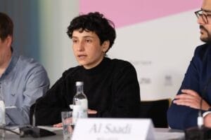 A woman with short curly hair speaks at a conference. She is wearing a black turtleneck sweater and sitting next to two men who are engrossed in a discussion. There are microphones and bottles of water on the table.