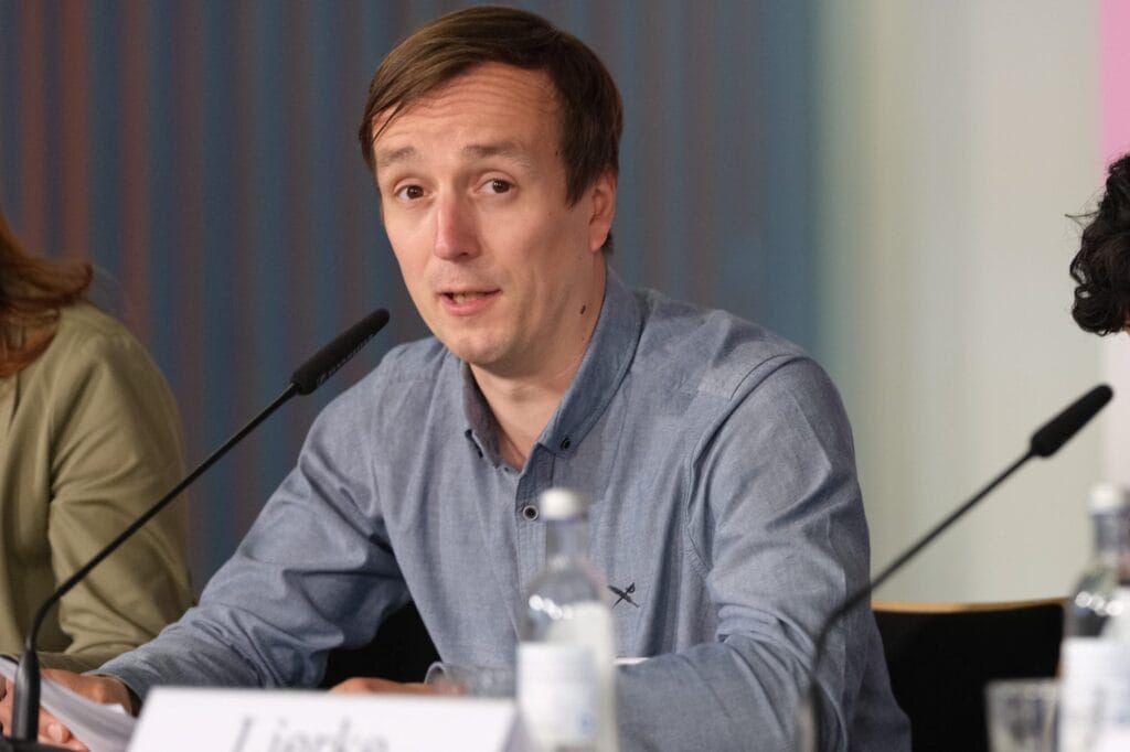 Ein Mann in einem blauen Hemd spricht auf einer Konferenz. Vor ihm sitzt ein Tisch mit Mikrofonen und einem Namensschild.