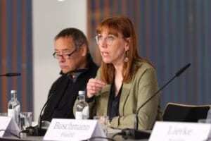 A woman with glasses speaking at a panel discussion sits next to a male panelist in a conference room with name badges and microphones.