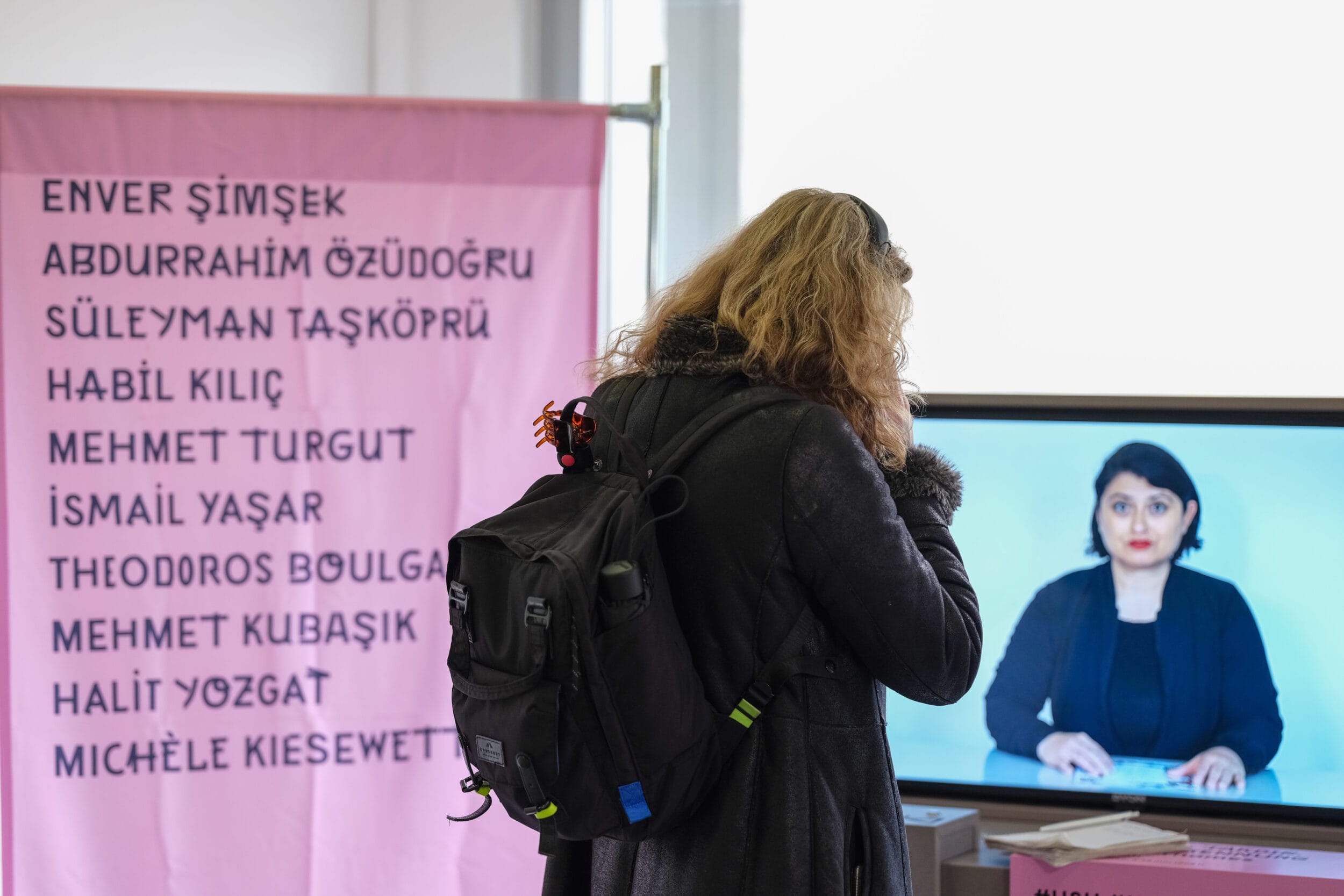 A woman with a rucksack looks at a memorial exhibition displayed on a screen with the names of individual people. Another woman can be seen on the screen.