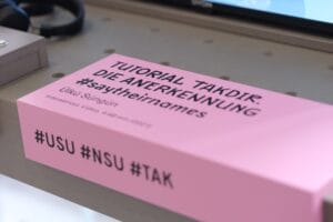 Rosafarbene Box mit der Aufschrift „#saytheirnames“ und „#usu“ auf einem Schreibtisch mit Kopfhörern im Hintergrund.