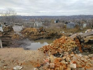 A destroyed urban area with scattered bricks and debris, a flooded crater, and distant buildings under a cloudy sky.