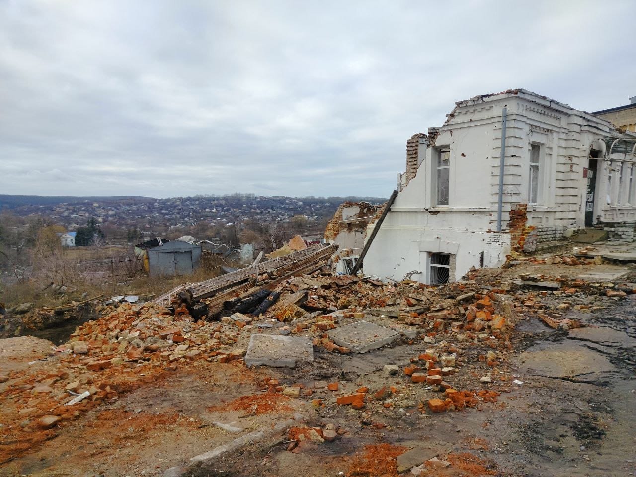 A partially collapsed building with scattered bricks and rubble in the foreground, overlooking a distant landscape under a cloudy sky.