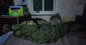 A military sleeping bag and gear scattered in a dimly lit room with a small window and cluttered table.