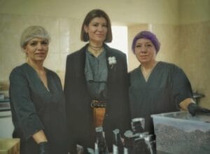 Three women standing together in a workspace: two in protective hairnets and gloves, one in a black outfit with a choker necklace. Surrounded by containers, these Ukrainian volunteers appear dedicated to their mission, possibly preparing to deliver food to those in need, even in the trenches.