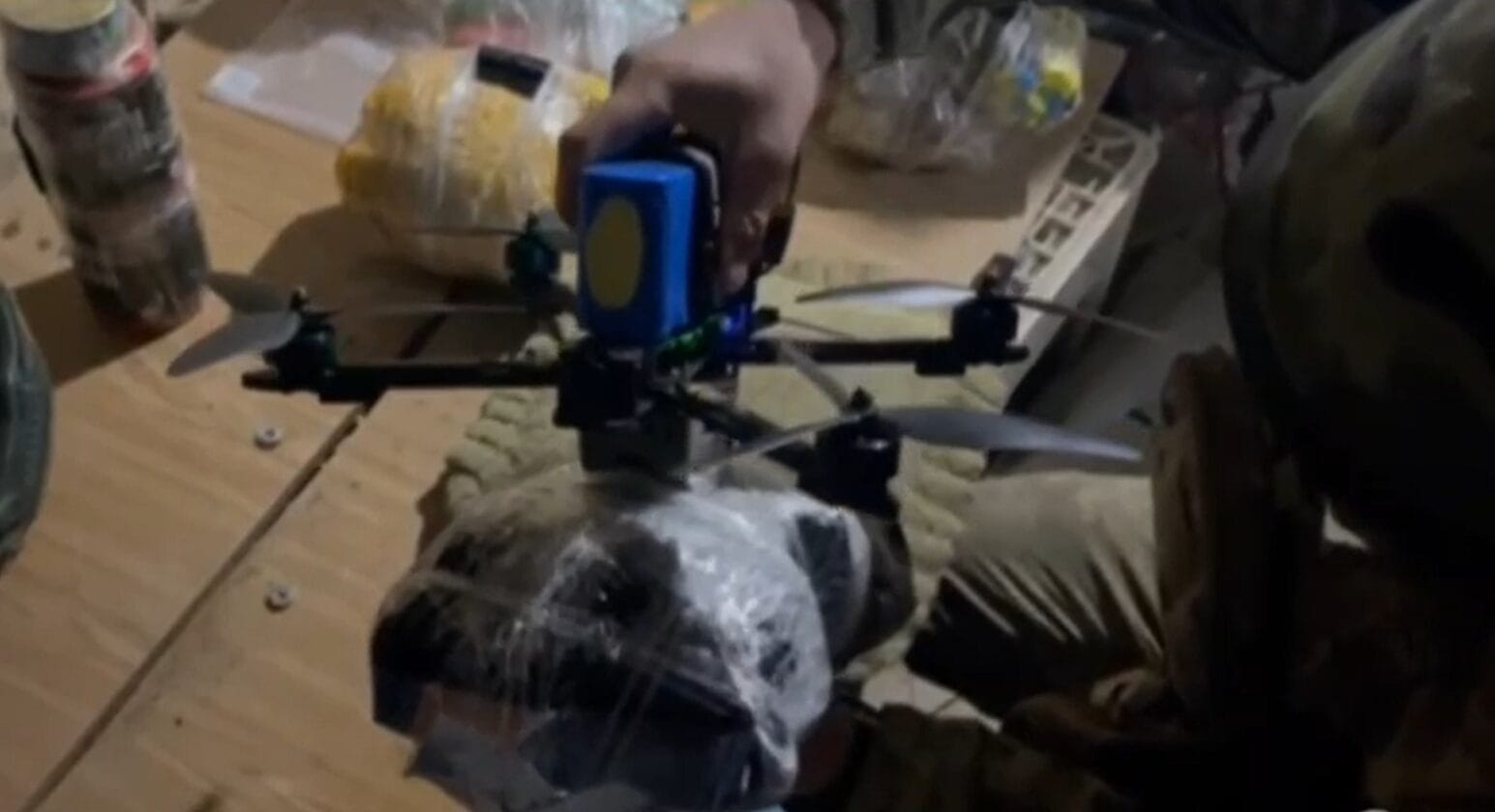 Person in camouflage clothing attaching supplies to a small drone on a wooden surface.