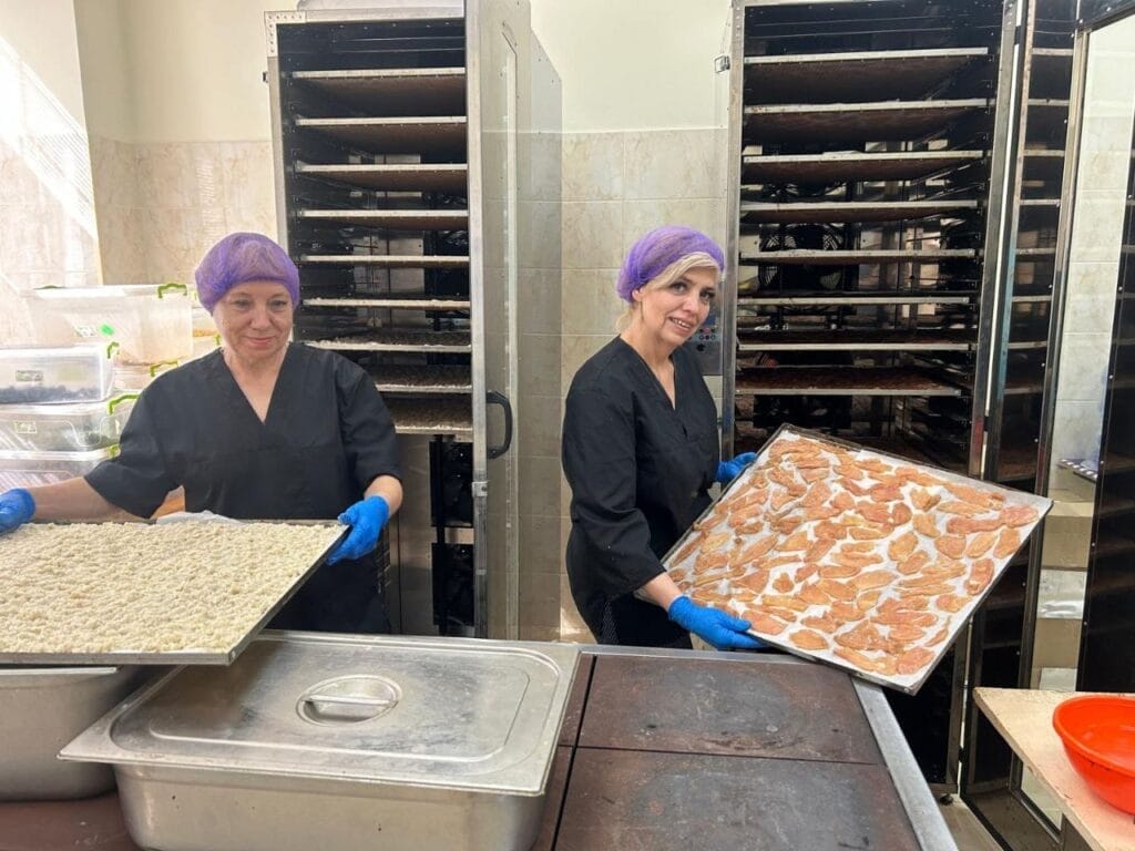 Two Ukrainian volunteers wearing black uniforms, blue gloves, and purple hair nets hold trays of food in a commercial kitchen with multiple ovens, preparing meals for food delivery to the trenches.