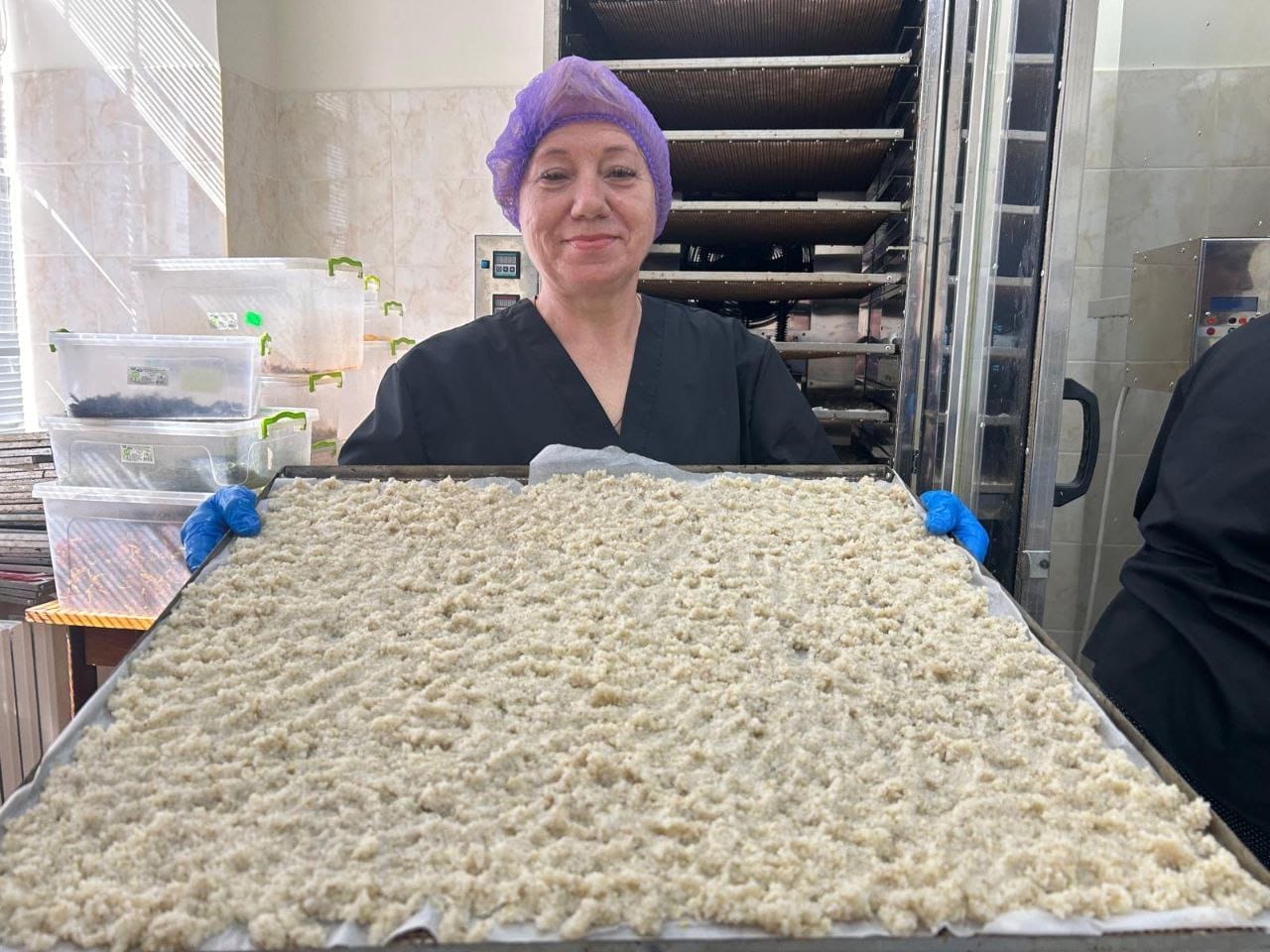 A person wearing a hairnet and gloves holds a large tray of uncooked shredded material, possibly food, in a kitchen with plastic bins and an industrial oven in the background, preparing for food delivery organized by Ukrainian volunteers.