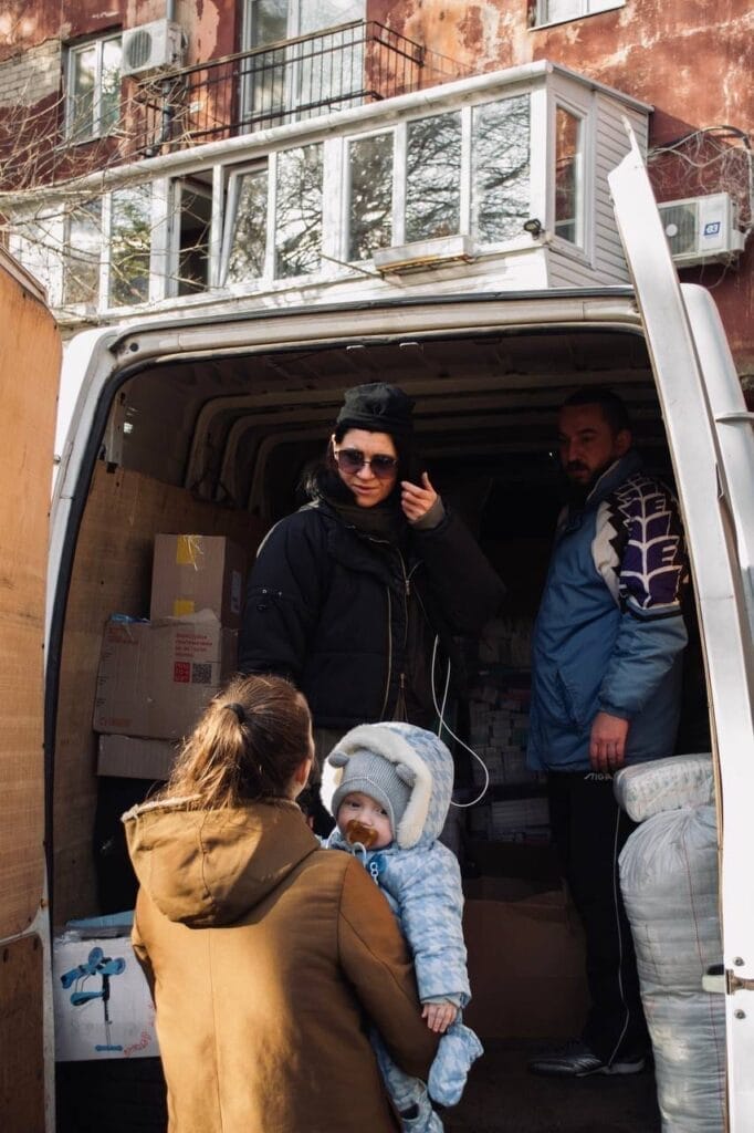 A woman in a black jacket stands inside a van with another man, engaging with a woman holding a warmly clothed baby outside. Various boxes, likely containing food deliveries from Ukrainian volunteers, are visible inside the van.