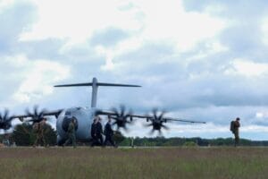 Ein großes Militärflugzeug steht mit laufenden Triebwerken auf einer Landebahn. Mehrere Personen gehen in der Nähe des Flugzeugs und ein Soldat läuft allein auf dem Gras rechts. Der Himmel ist bewölkt.