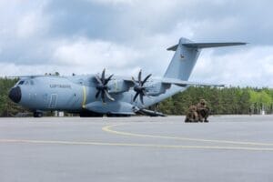 Ein Militärtransportflugzeug mit der Aufschrift „Luftwaffe“ auf der Seite steht auf einer Startbahn. Daneben stehen zwei Militärangehörige und zwei Militärhunde. Im Hintergrund sind Bäume zu sehen.