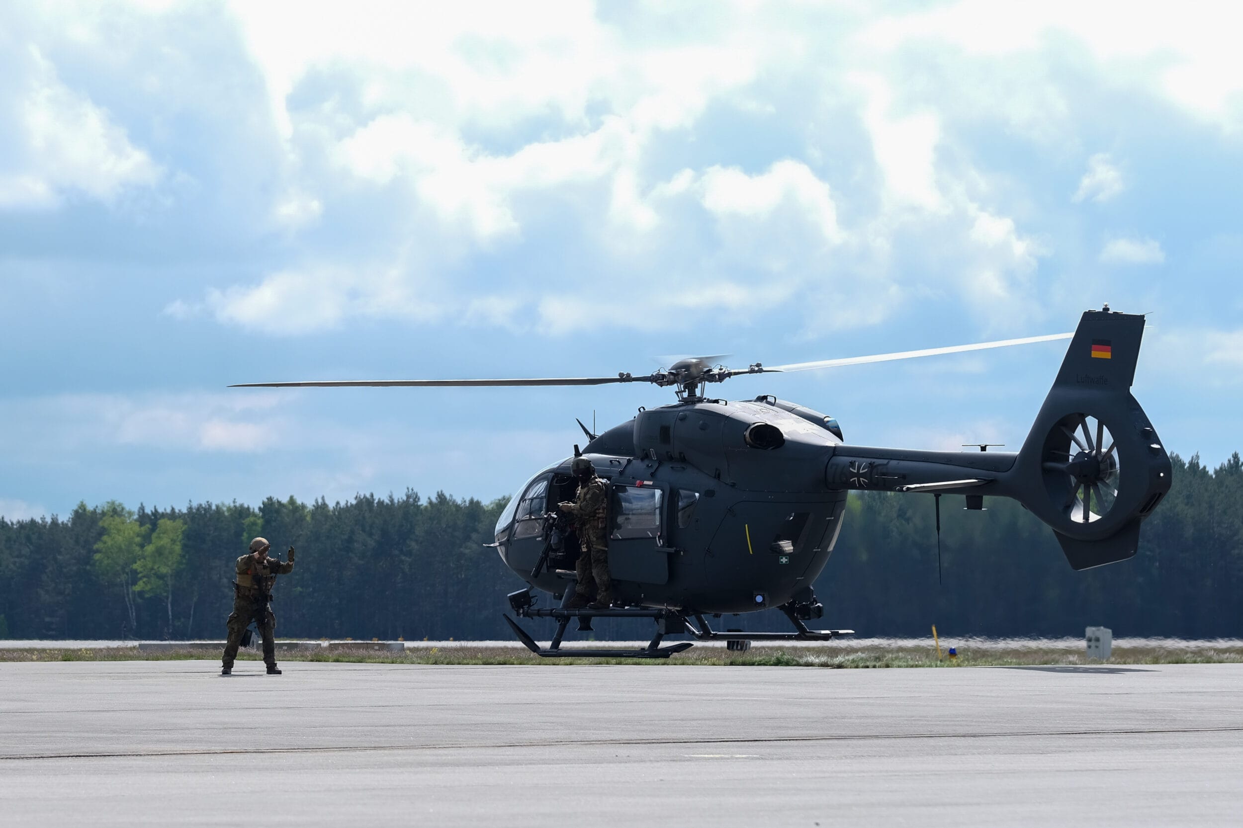 Ein Militärhubschrauber auf einer Landebahn. Ein Soldat gestikuliert, ein anderer sitzt an der offenen Seitentür. Der Himmel ist teilweise bewölkt und im Hintergrund ist eine Baumreihe zu sehen.