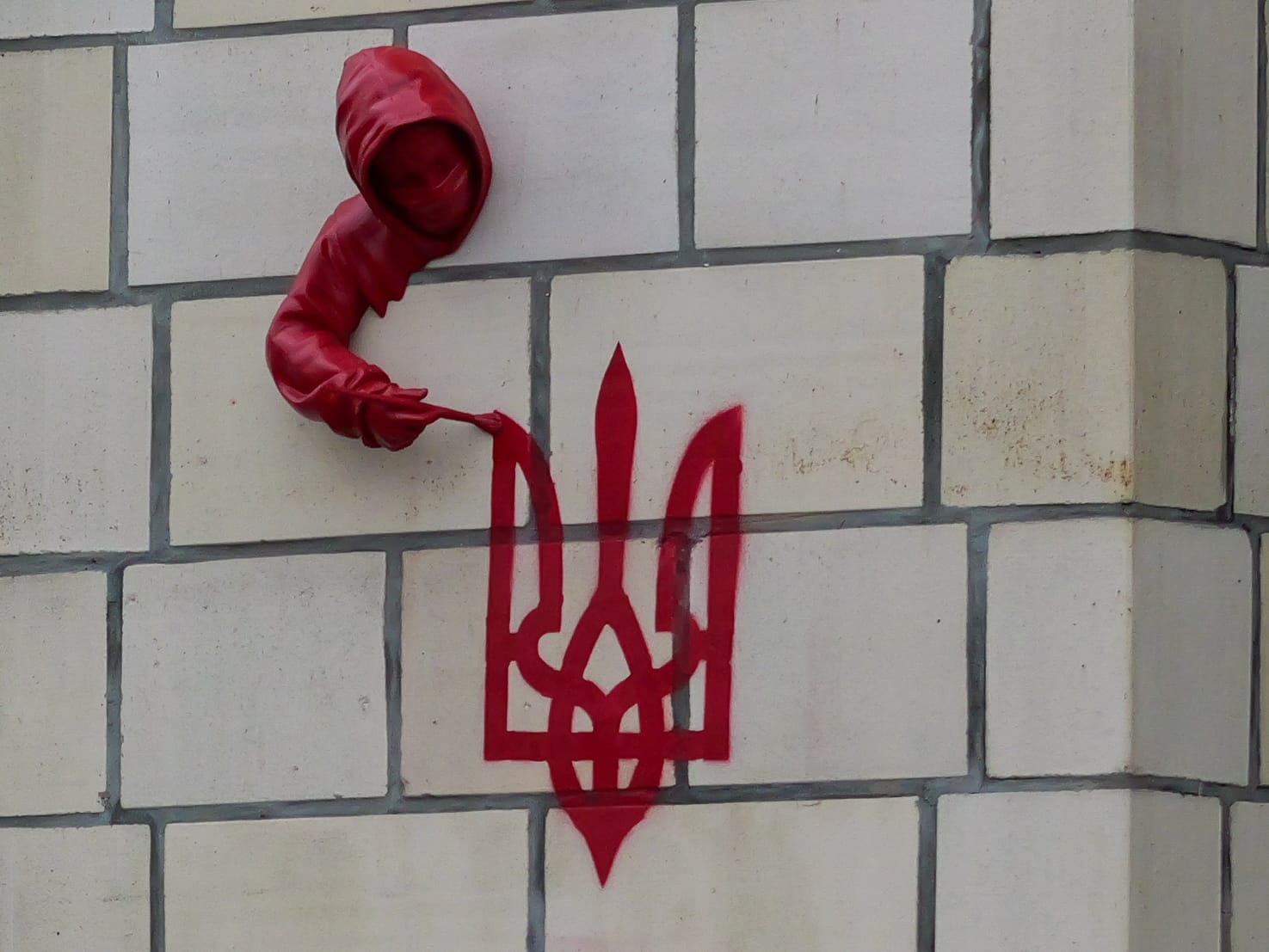 Aus einer Wand ragt die rote Skulptur einer vermummten Gestalt und zeichnet das Wappen der Ukraine in roter Farbe.