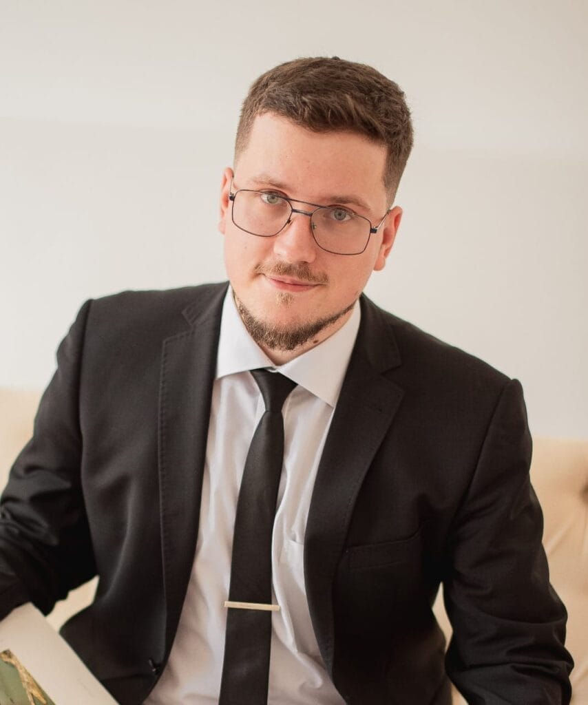 Oleksandr Kraiev wears a black suit and tie and sits there holding a book in his hand. He wears glasses, has short hair and a trimmed beard. 