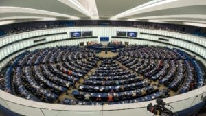 Das EU-Parlament in Straßburg ist ein großer, runder Saal mit zahlreichen Menschen an Schreibtischen, die einem zentralen Podium gegenübersitzen. Mehrere Flaggen werden gezeigt und auf digitalen Bildschirmen werden Präsentationen gezeigt.