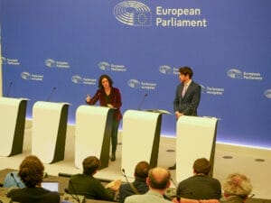 Two people stand with microphones at speakers and address an attentive audience in a conference room decorated with the EU Parliament logo.