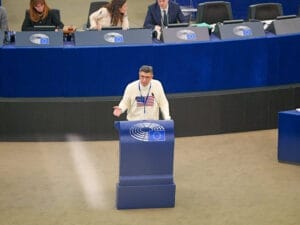 A person speaks at a podium in a large meeting room with blue desks, highlighted by the flags and emblem of the EU Parliament.