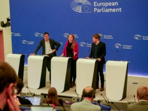 Three people stand behind speakers in a conference room of the European Parliament and speak to an audience. The logo and text of the EU Parliament are clearly visible on the blue background.
