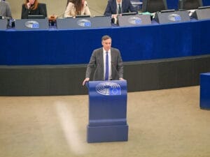 A man in a suit speaks at a podium in the large assembly hall of the EU Parliament, recognizable by its blue seats and the insignia of the European Union.