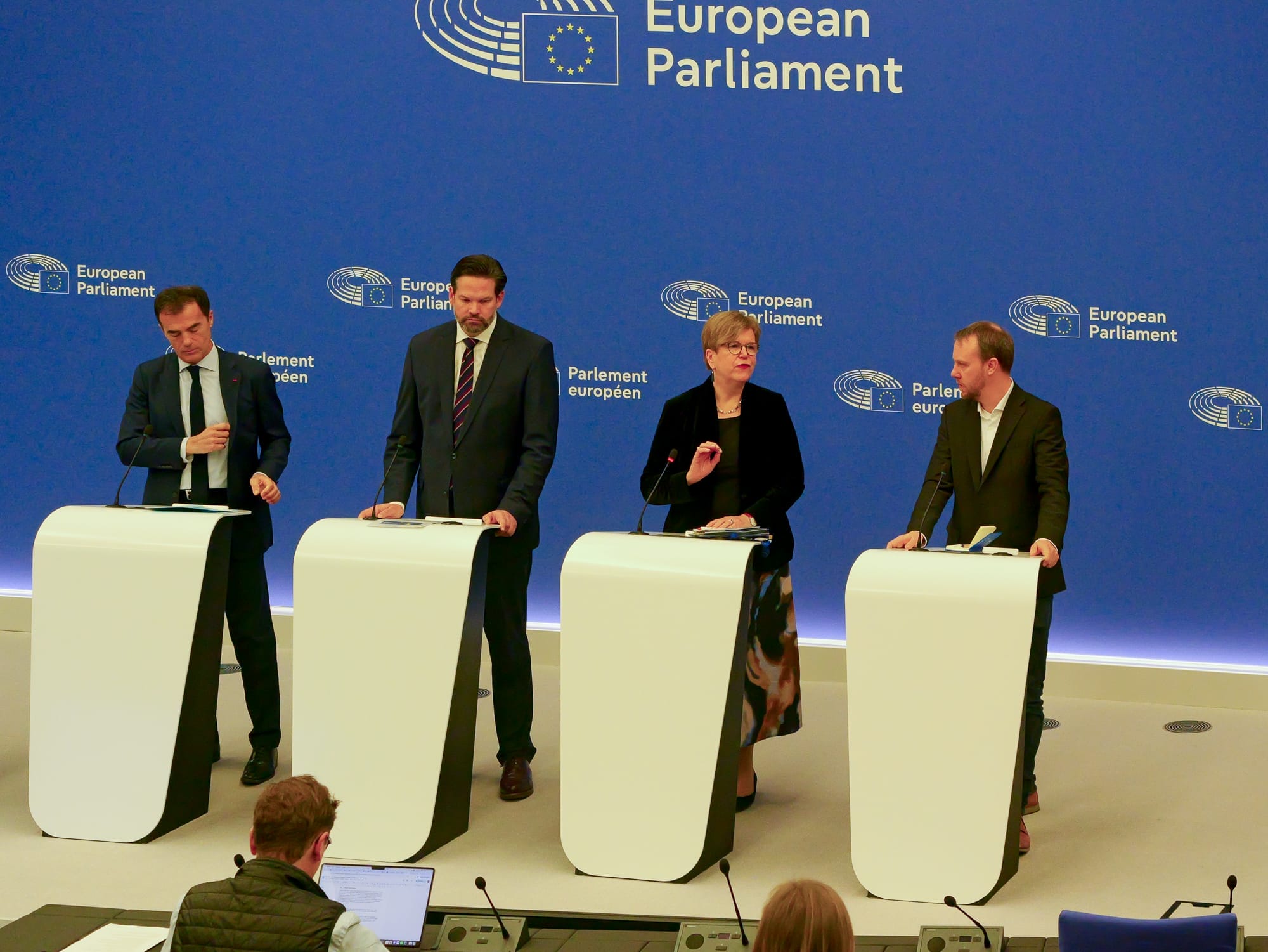 At a European Parliament press conference, four people stand at lecterns, speaking and gesticulating. A spectator can be seen in the foreground. 