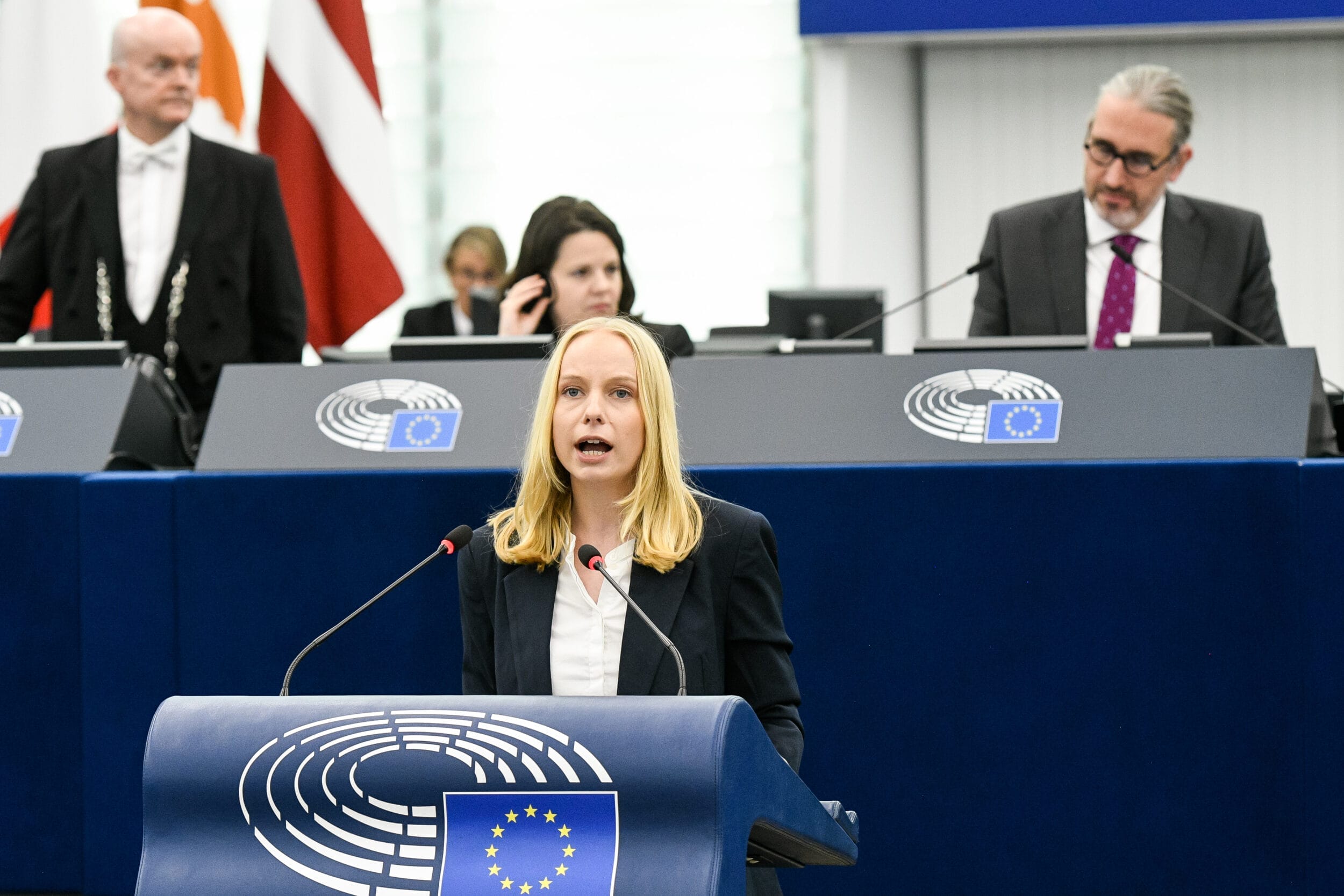 Sabrina Repp hält eine Rede vor dem Publikum an einem mit dem Emblem des Europäischen Parlaments geschmückten Podium in Straßburg und zieht die Aufmerksamkeit der anderen auf sich, die gespannt zuhören.