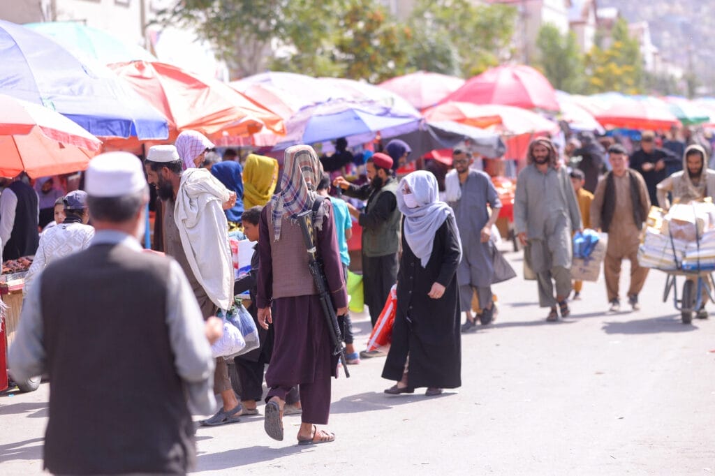 A crowded outdoor market in Kabul, Afghanistan, with colorful umbrellas and passers-by. One person carries a weapon, another pushes a cart loaded with crates.