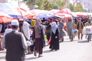 A crowded outdoor market in Kabul, Afghanistan, with colorful umbrellas and passers-by. One person carries a weapon, another pushes a cart loaded with crates. 