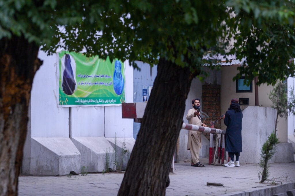 Banners with veiling rules for women dominated the streetscape. A man and a woman are standing at a barrier in front of a building with a green sign. Trees partially obscure the view.
